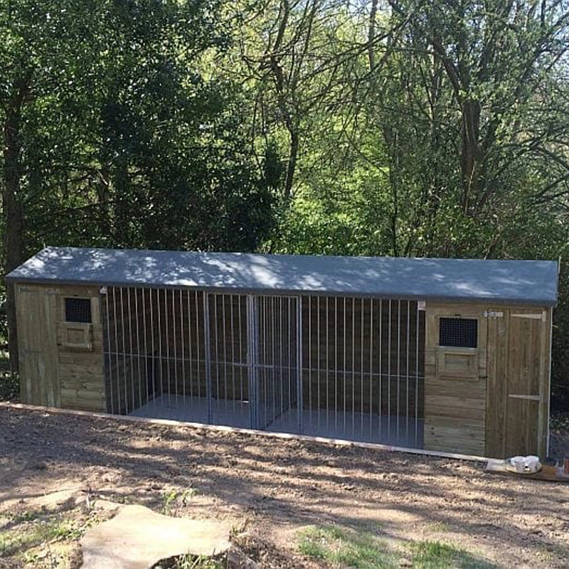 KENNELSTORE Wymbury Double Wooden Dog Kennel, a 18ft x 5ft multi-dog housing unit. The kennel features a distinctive apex roof and is made from pressure-treated tanalised timber, with two separate runs equipped with galvanised panels for security.